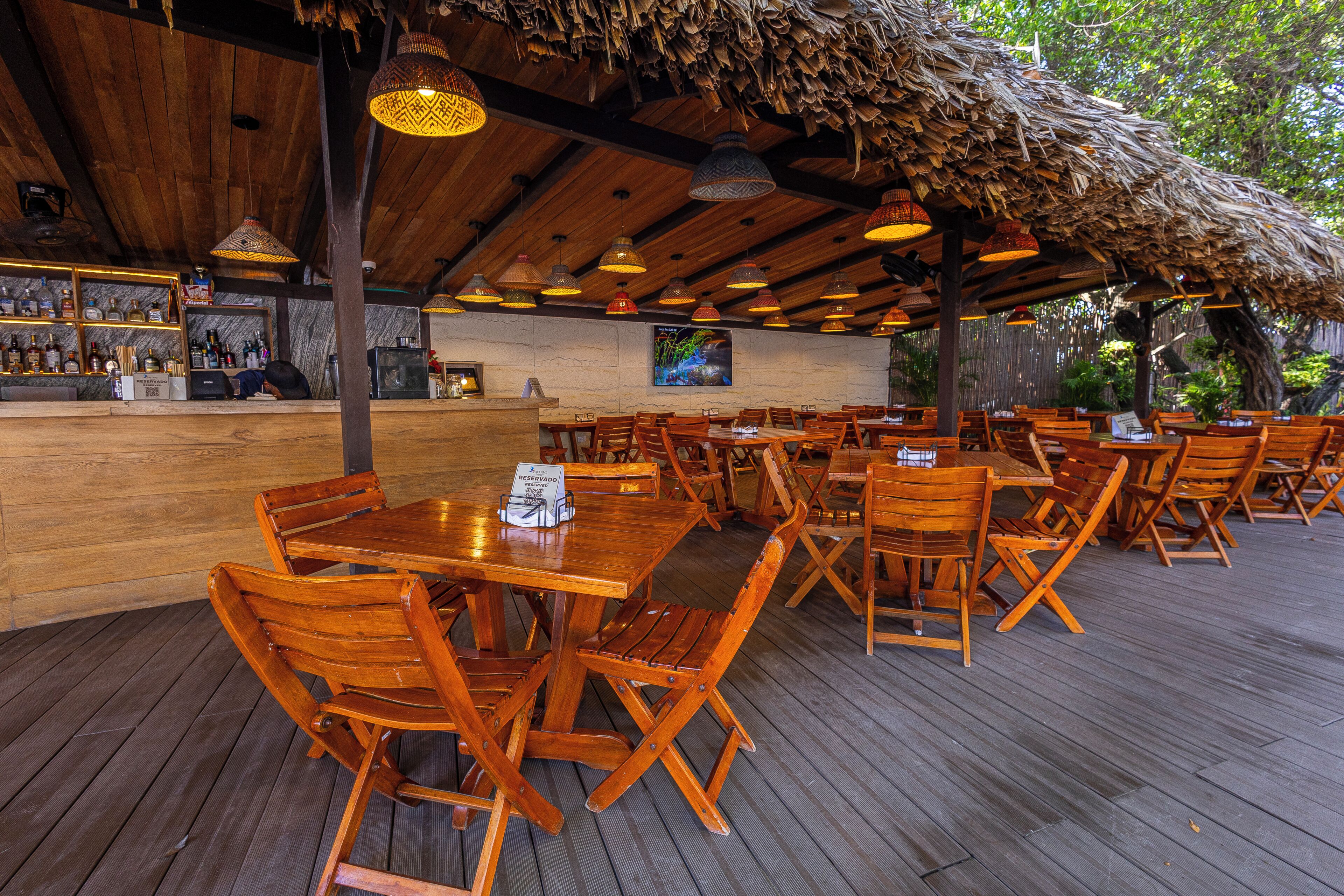 An outdoor restaurant with wooden tables and chairs, a bar area with bottles, and a thatched roof.