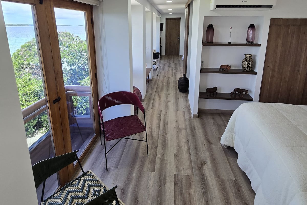 A hotel room with a bed, a chair, a shelf with decorative items, and a view of the outdoors.