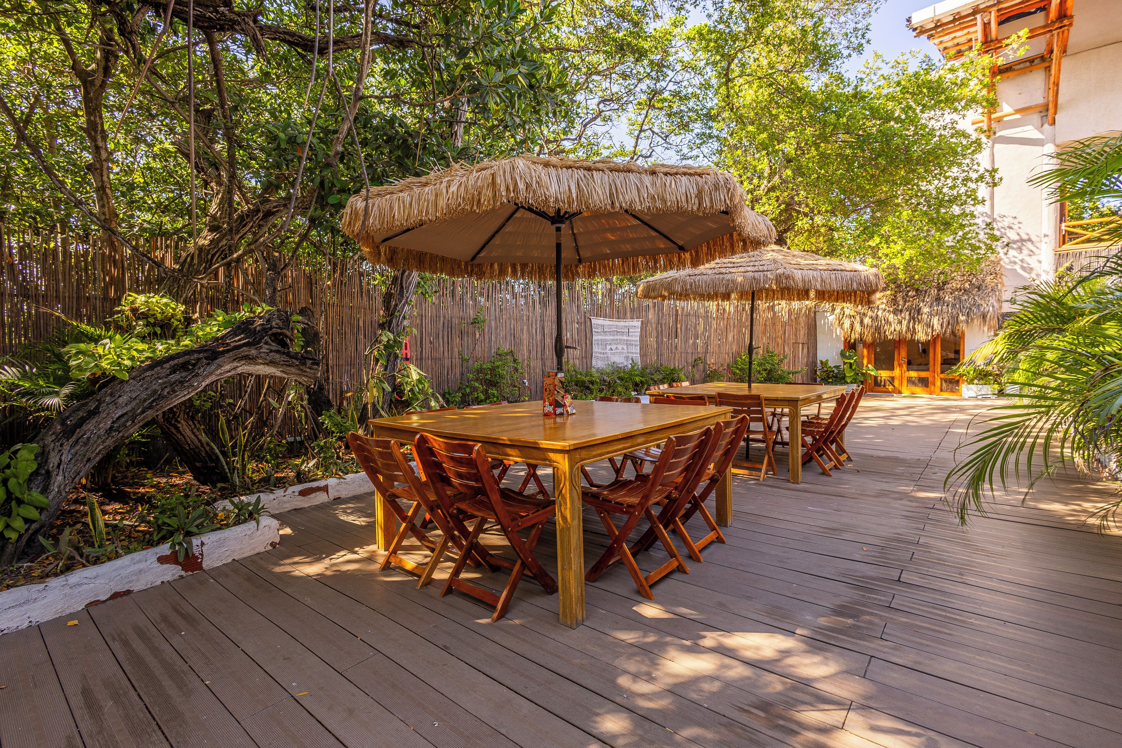 An outdoor dining area with wooden tables and chairs, surrounded by lush greenery and a thatched roof structure.