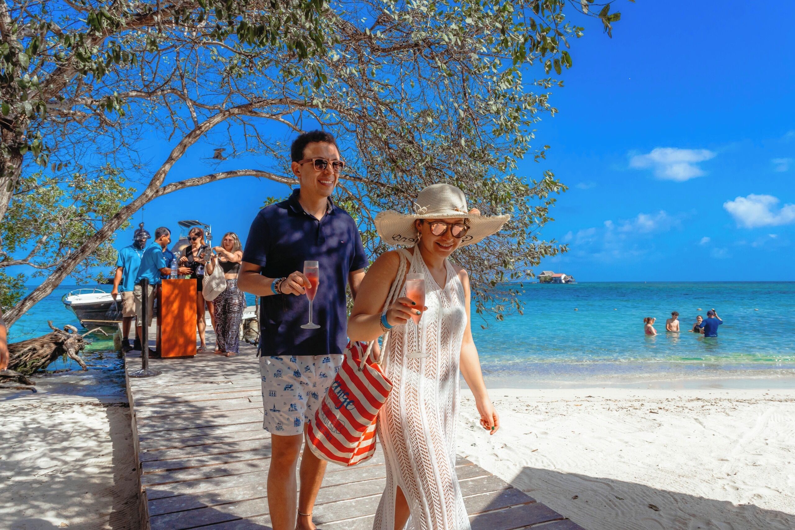 A wooden pier leading to a beach with clear blue water, a boat, and people enjoying the seaside.