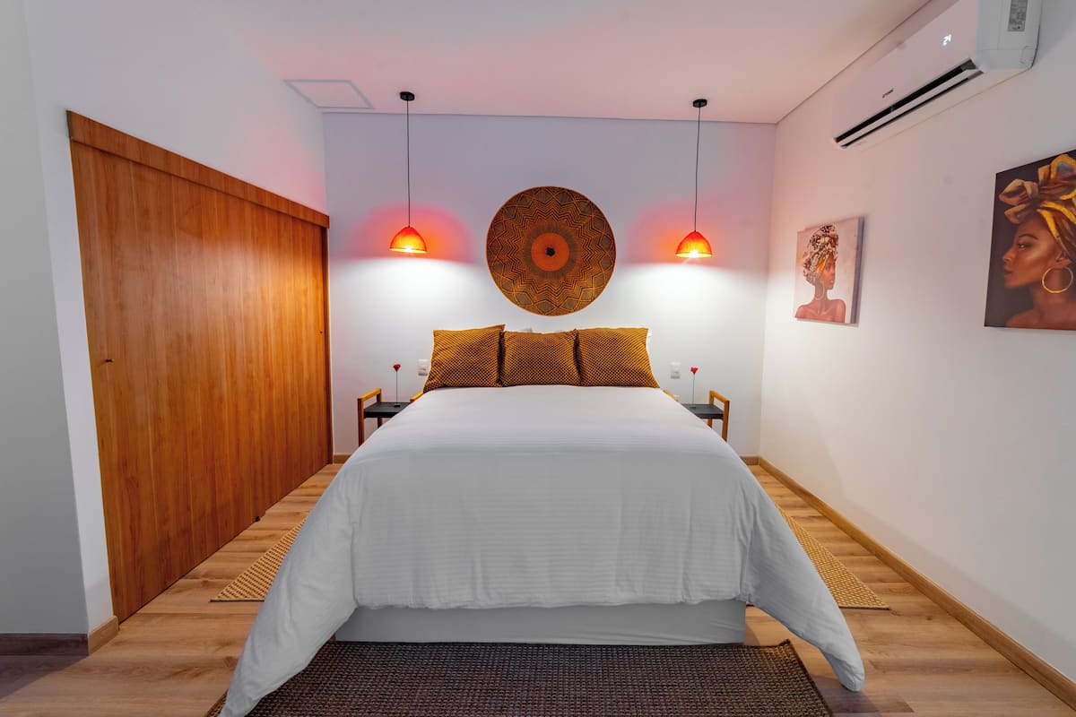 A bedroom with a wooden wardrobe, a bed with white bedding, two pendant lights, and a wall-mounted decorative plate.