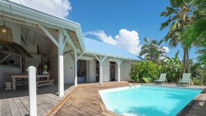 A wooden deck with a pool, white building with a blue roof, and palm trees. - Complete relaxation at Villa Lakaz with its spacious terrace and private pool. (Le Moule)