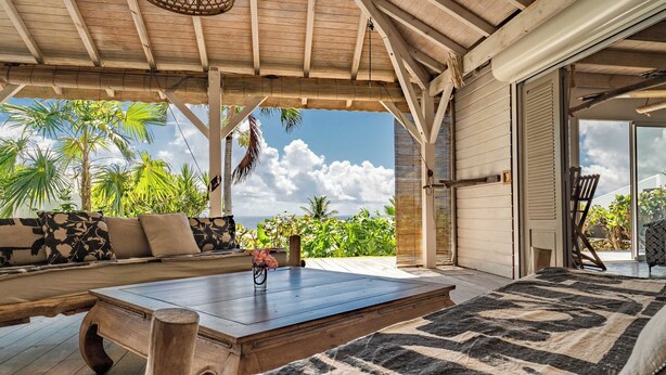 A covered outdoor seating area with a wooden table, a sofa with patterned cushions, and a view of the ocean and palm trees. - Complete relaxation at Villa Lakaz with its spacious terrace and private pool. (Le Moule)
