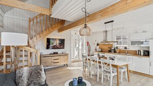 Una cocina moderna con armarios blancos y detalles de madera, un comedor con sillas blancas y una escalera con vigas de madera a la vista.