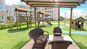 A children's play area with a slide, swings, and a small wooden hut. There are wicker chairs and a pergola overhead.