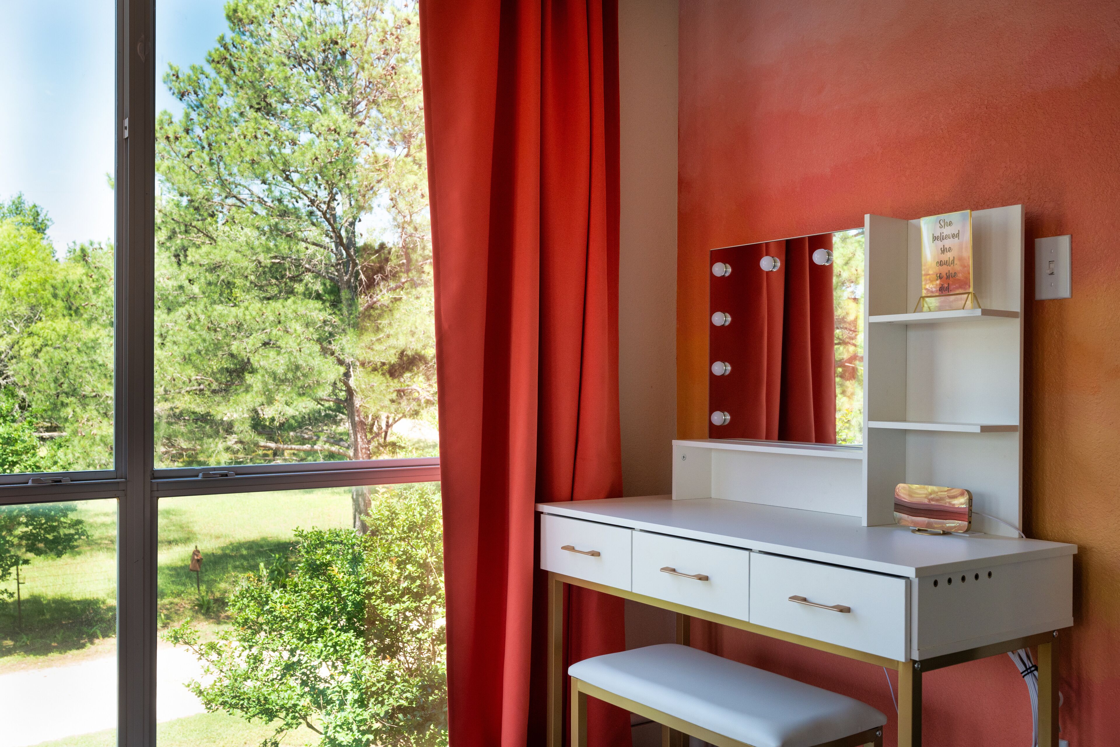 Habitación con escritorio, espejo y banco. Ventana con cortinas rojas y vista a zonas verdes.