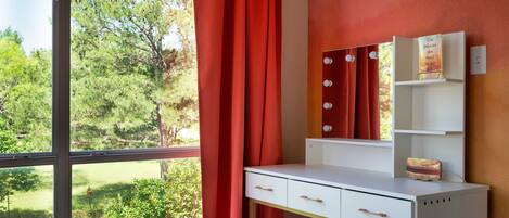 Habitación con escritorio, espejo y banco. Ventana con cortinas rojas y vista a zonas verdes.