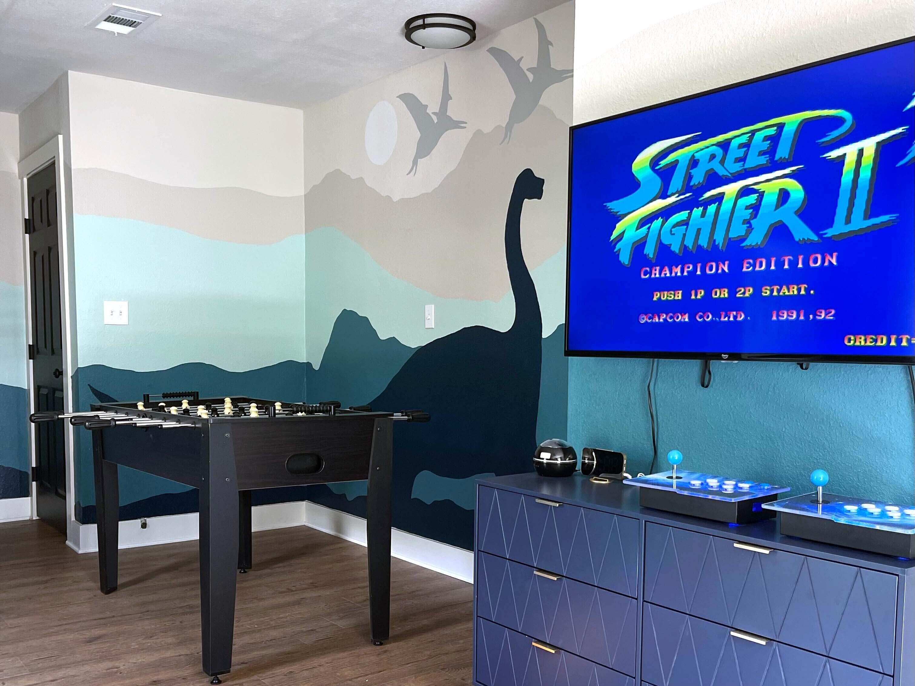 A room with a foosball table, a TV displaying 'Street Fighter II Champion Edition', and a wall with a mountain and bird mural.