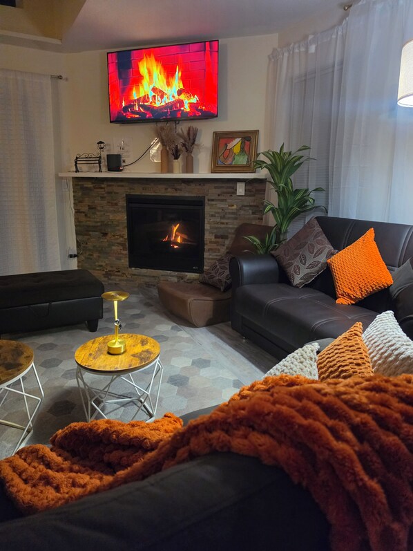 A living room with a fireplace, a television mounted above it, a sofa with cushions, and a coffee table. - New Arcades Game Room, Firepit, Pet Friendly (Hamlin)