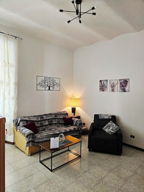 A living room with a sofa, a coffee table, a lamp, and a chair. There are wall art pieces and a ceiling light fixture.