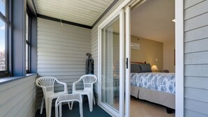 Un balcon avec deux chaises en plastique blanc, une porte-fenêtre coulissante donnant sur une chambre avec un lit et une table de chevet, et une fenêtre avec des stores.