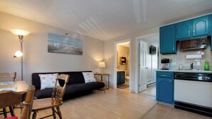 Une cuisine moderne avec des armoires de couleur turquoise, un lave-vaisselle et un four à micro-ondes. On y trouve un coin repas avec des chaises et une table en bois, ainsi qu’un salon avec un canapé noir et un tableau au mur.