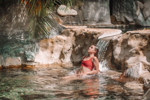 A person in a red swimsuit in a natural water setting with rocky formations and a palm tree. - Thermomania (Fortuna)