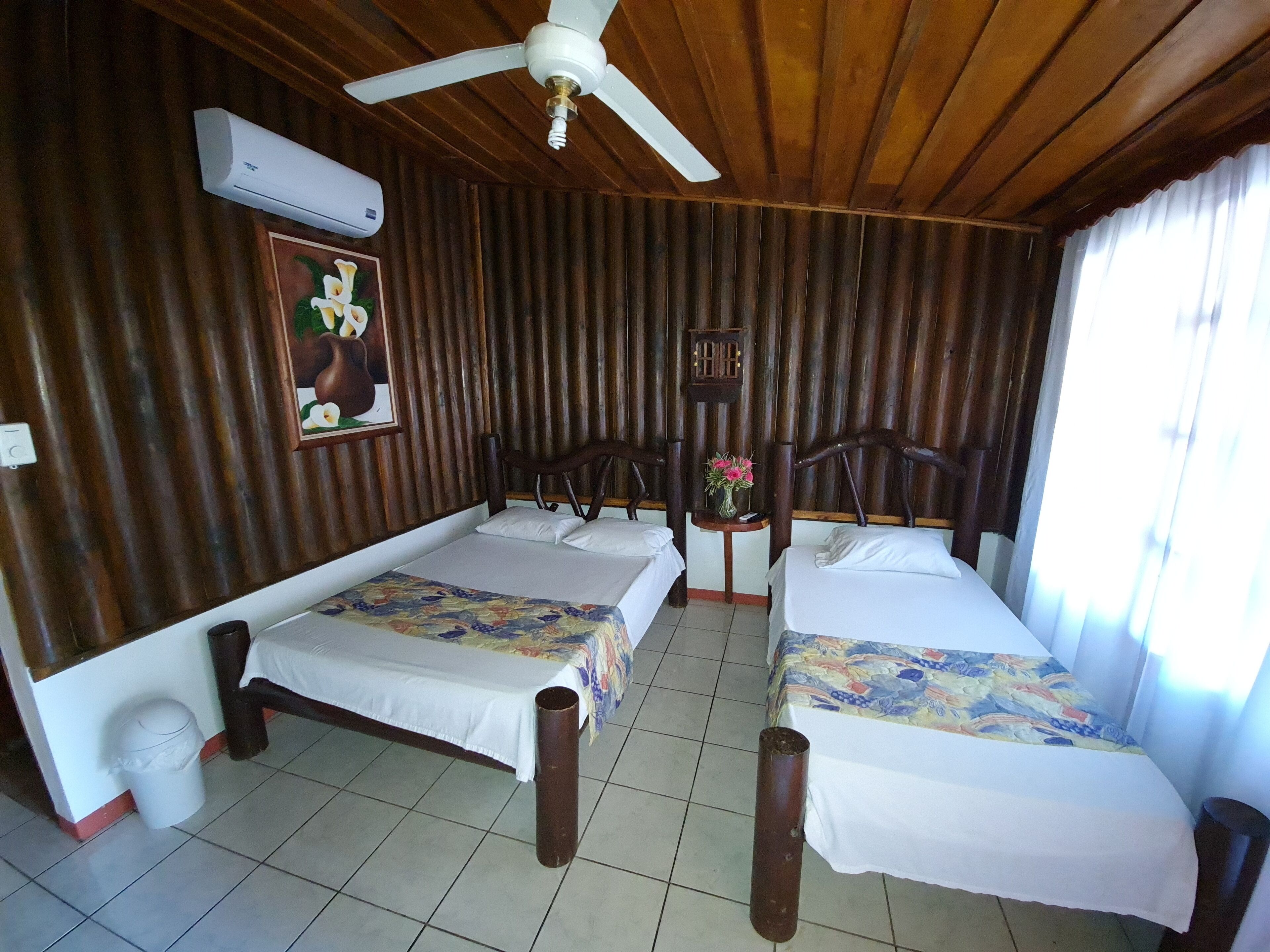 A room with two beds, a ceiling fan, an air conditioning unit, a small table with a vase of flowers, and a wall-mounted clock.