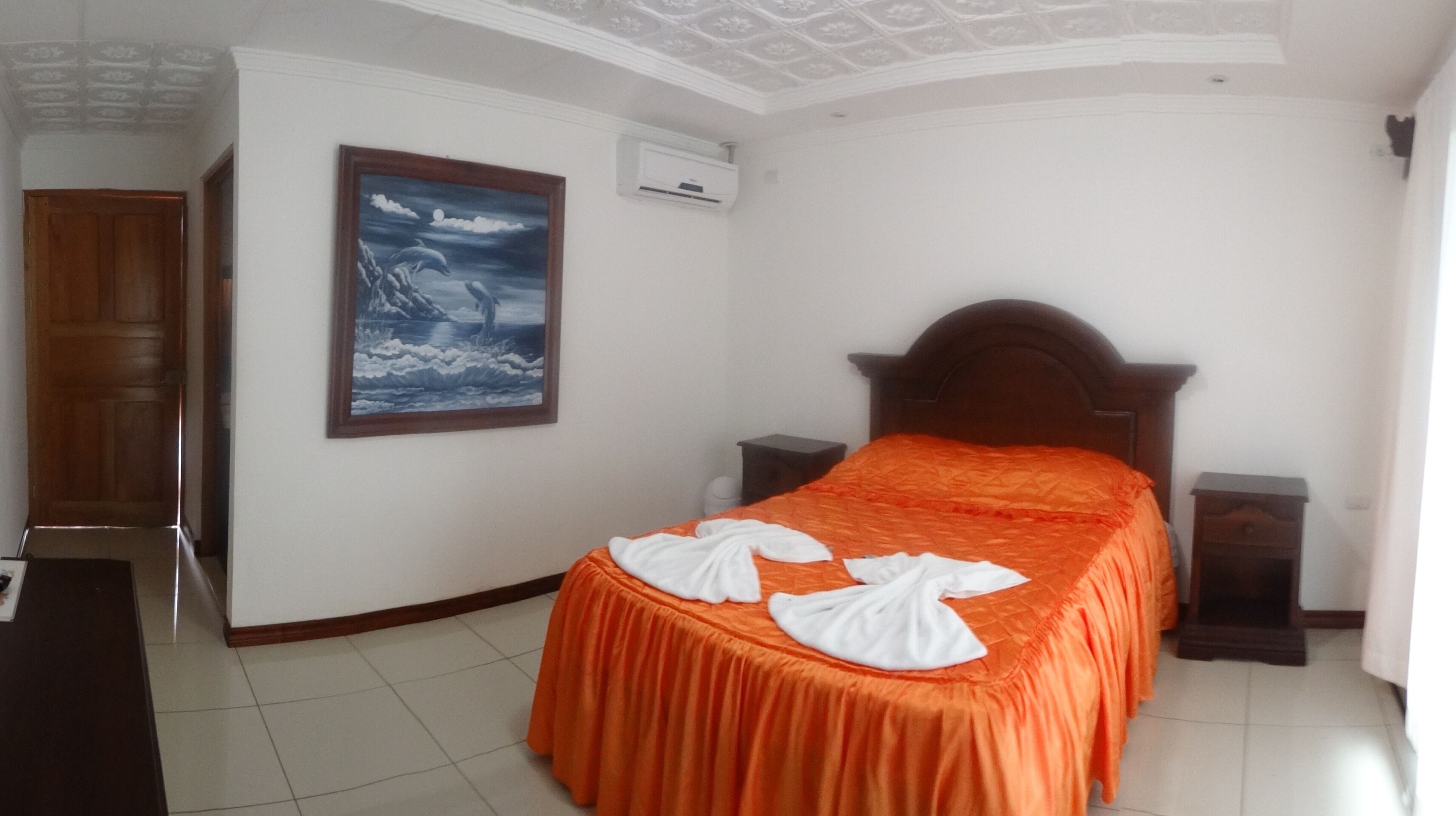 A hotel room with a bed, a wooden headboard, a nightstand, a painting on the wall, and an air conditioning unit.