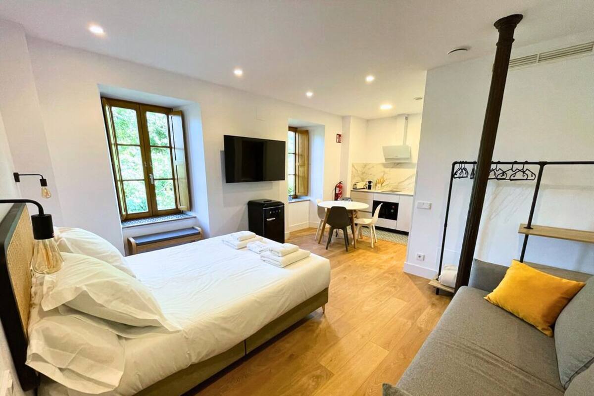 A modern hotel room with a large bed, a sofa, a dining area, and a kitchen.