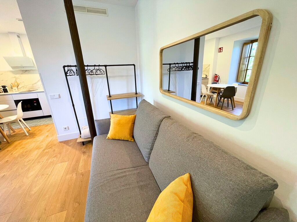 Un salon moderne avec un canapé gris, des coussins jaunes, un plancher en bois, un coin cuisine avec une cuisinière et un grand miroir au mur.