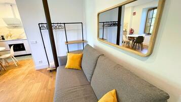 Un salon moderne avec un canapé gris, des coussins jaunes, un plancher en bois, un coin cuisine avec une cuisinière et un grand miroir au mur.