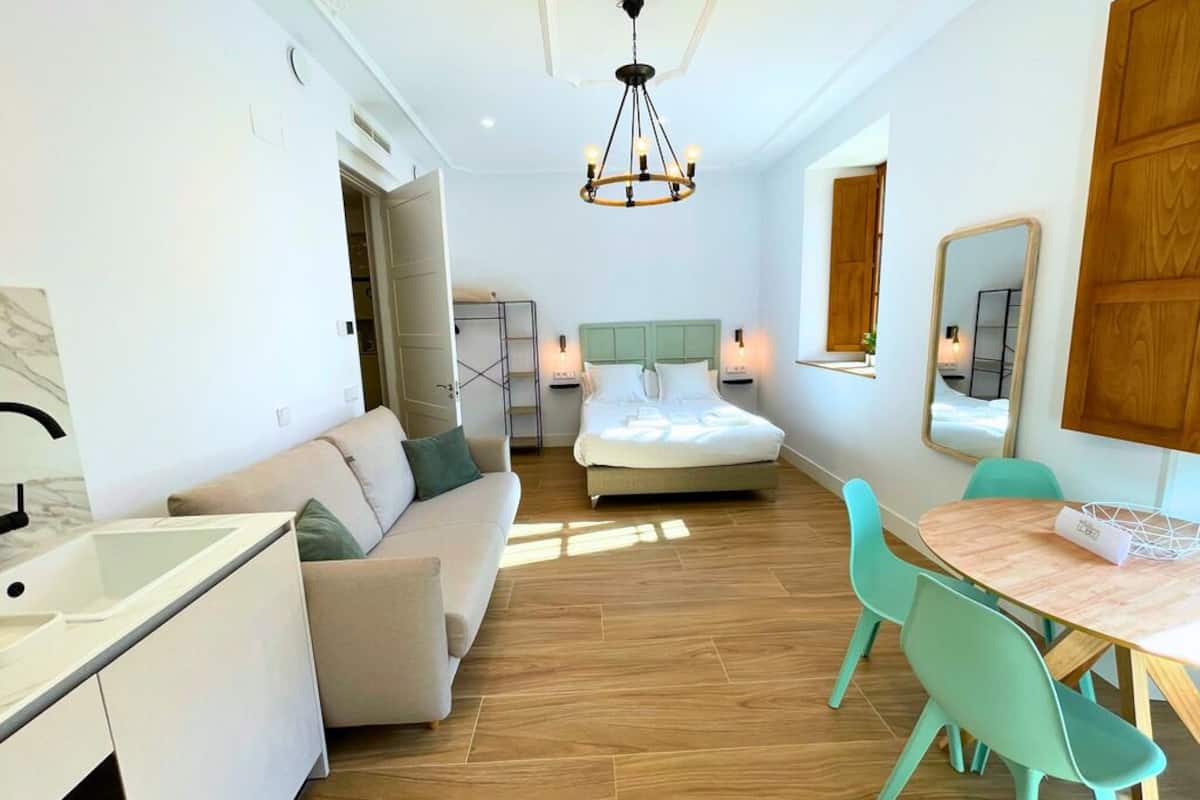 A modern hotel room with a bed, a sofa, a dining table with chairs, a kitchen area, and a large mirror.