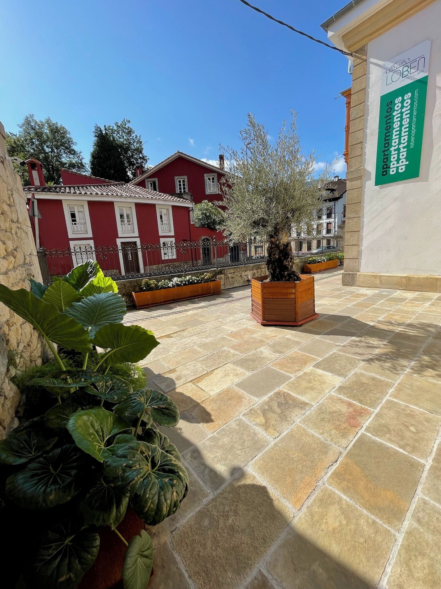 A courtyard with a red building, a wooden planter with a tree, and a sign for 'apartamentos'.
