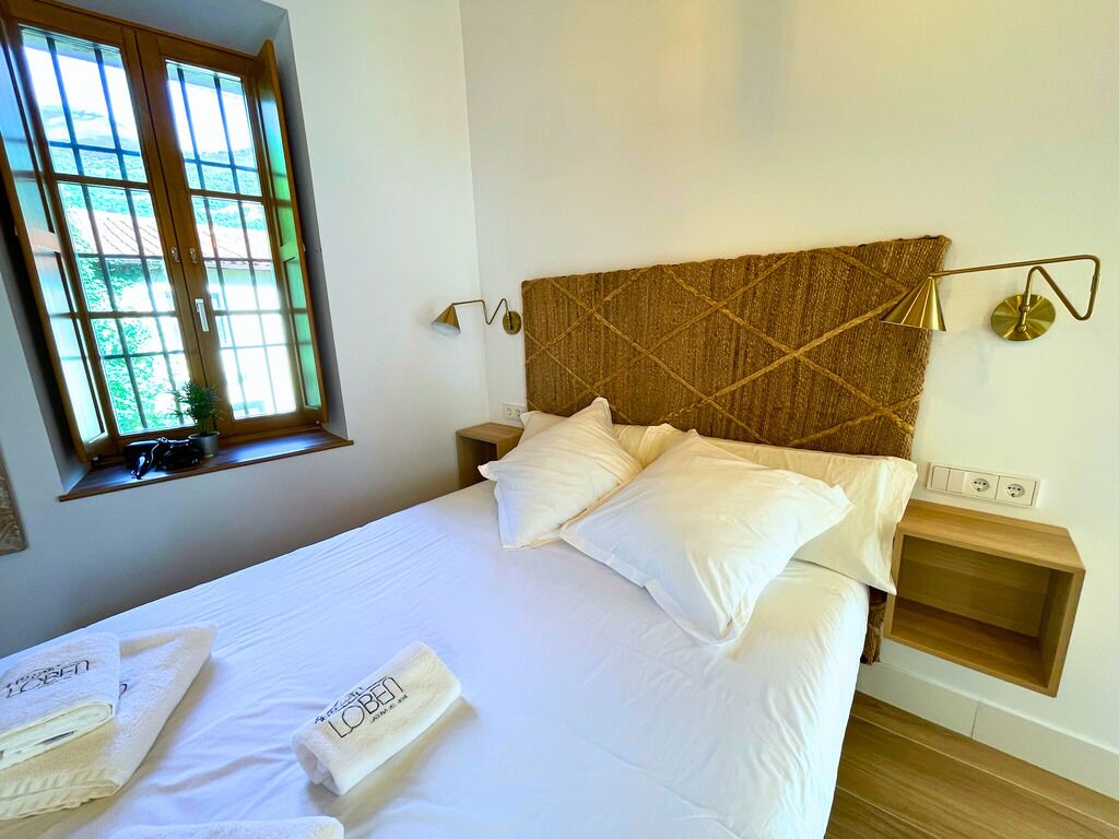 Una cama bien hecha con sábanas y almohadas blancas, cabecera de madera, mesita de noche con lámpara y ventana con vistas a zonas verdes.