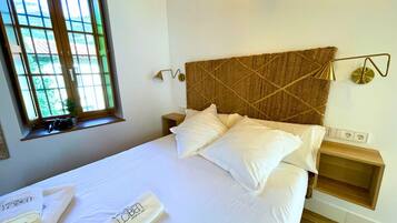 Una cama bien hecha con sábanas y almohadas blancas, cabecera de madera, mesita de noche con lámpara y ventana con vistas a zonas verdes.