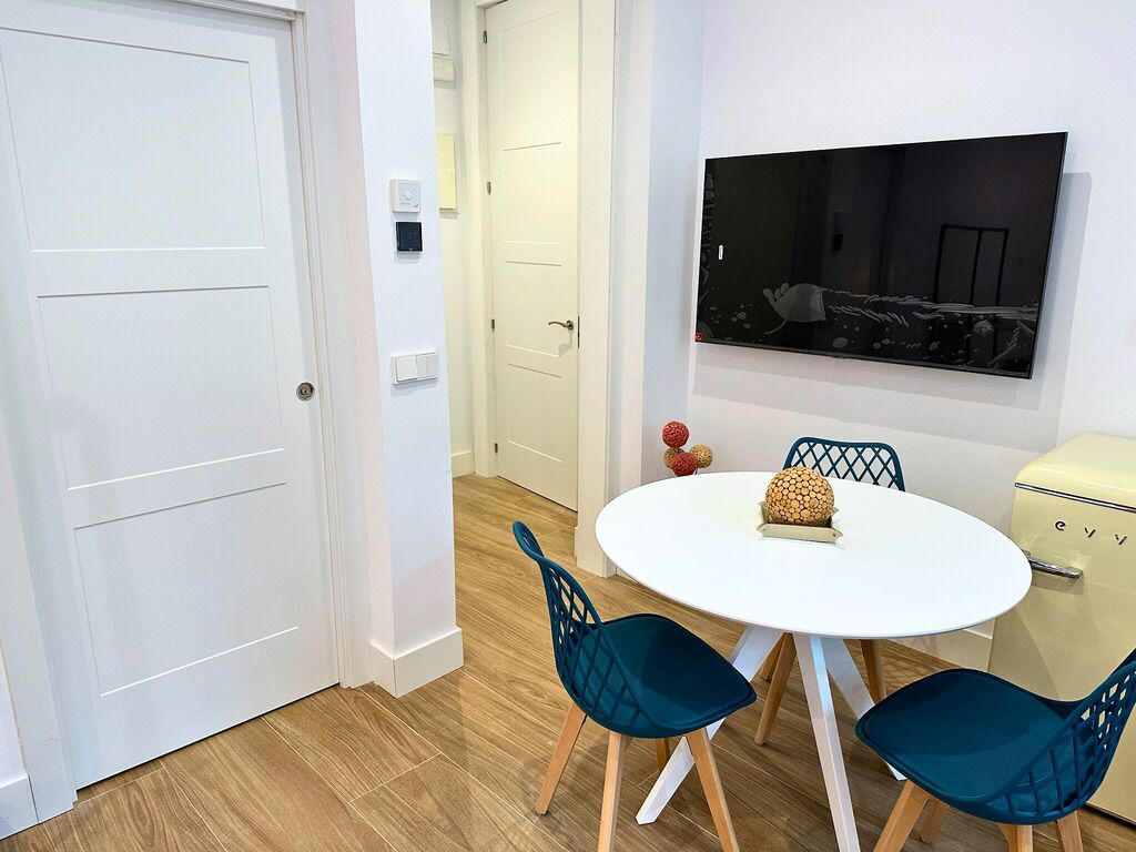 Un coin repas avec une table blanche, des chaises de couleur turquoise, un téléviseur fixé au mur et un réfrigérateur de style rétro.