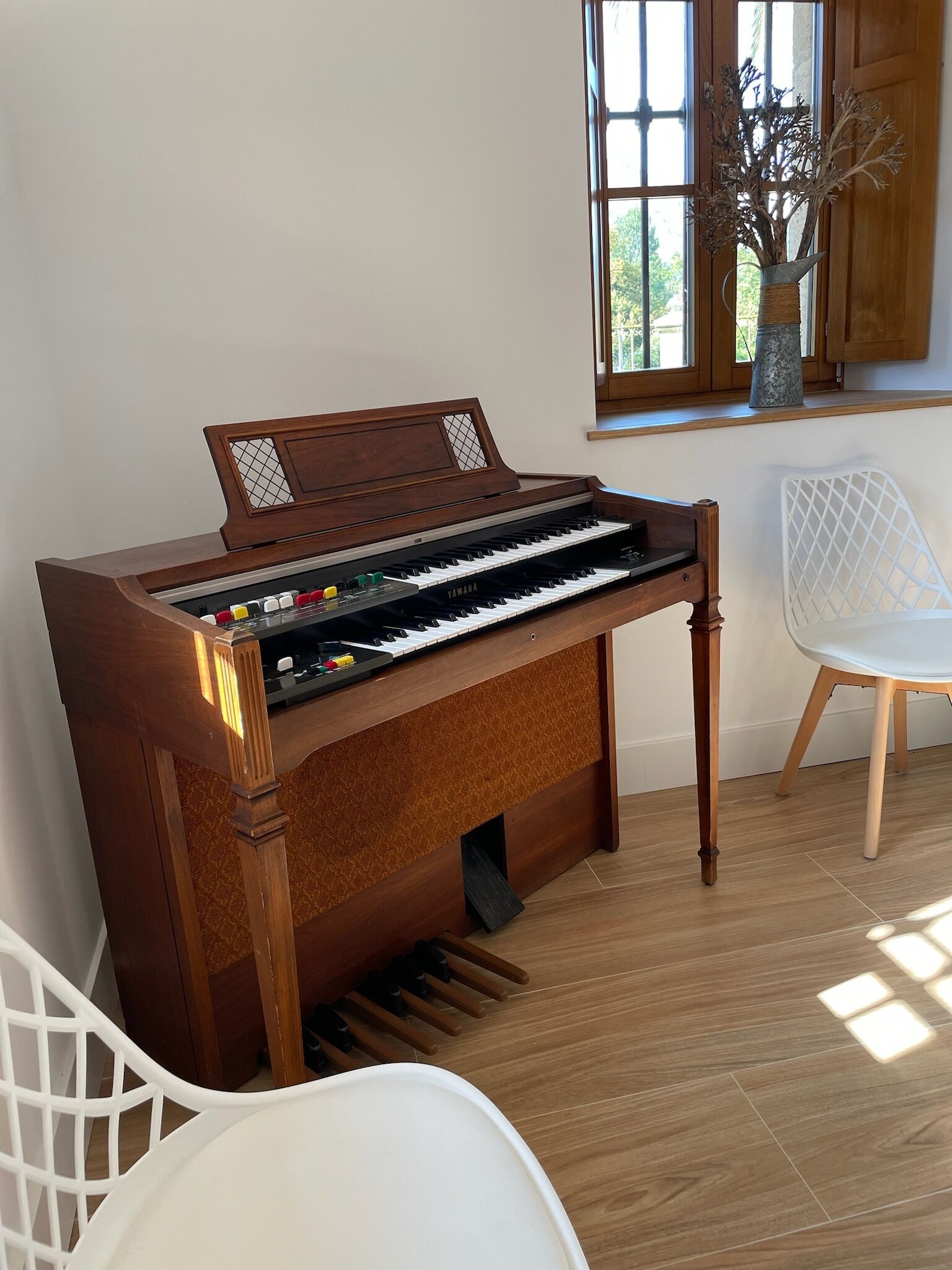 Un piano en una habitación con una ventana, una silla blanca y un florero con ramas.