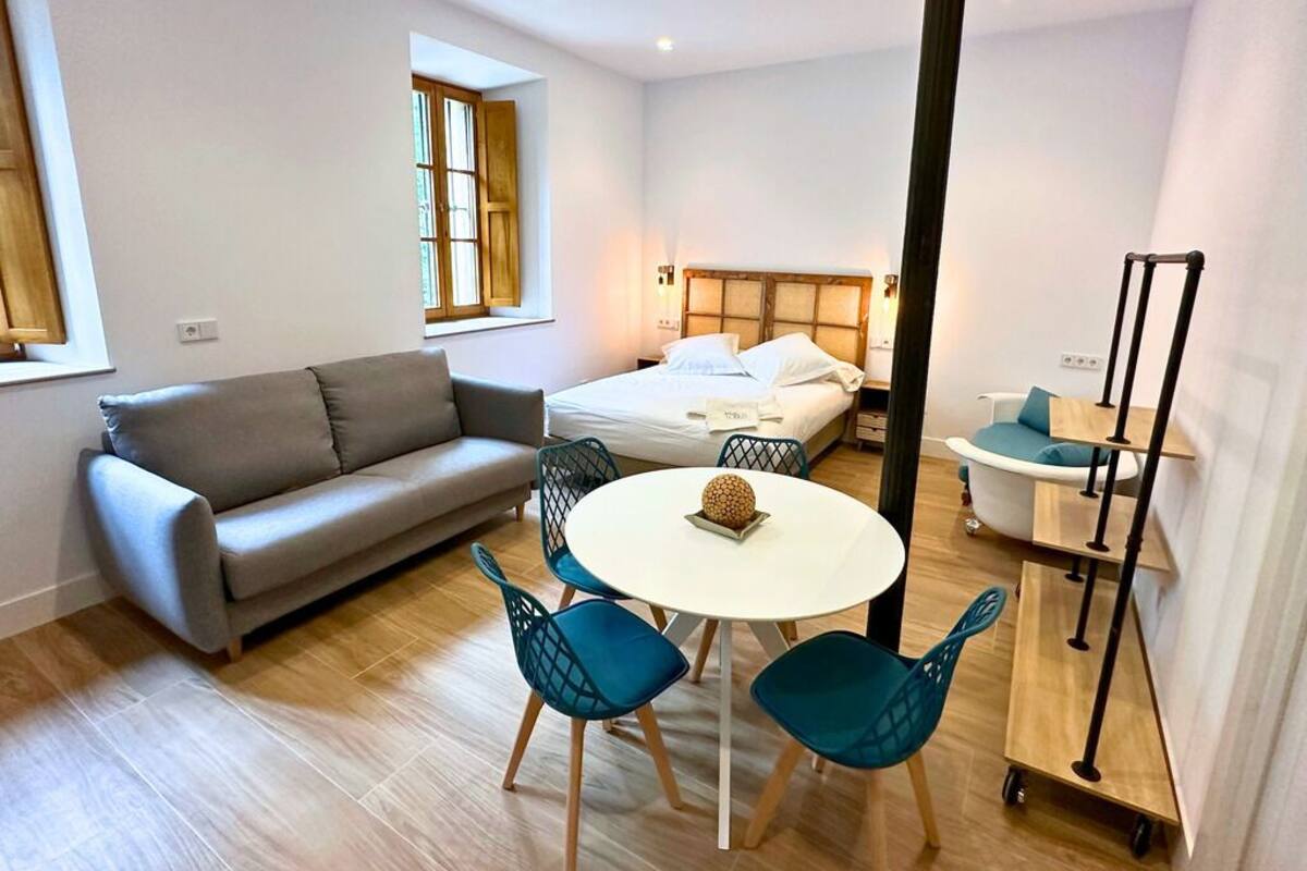 A modern, well-lit room with a sofa, a round table with chairs, a bed with a wooden headboard, and a small kitchenette area.