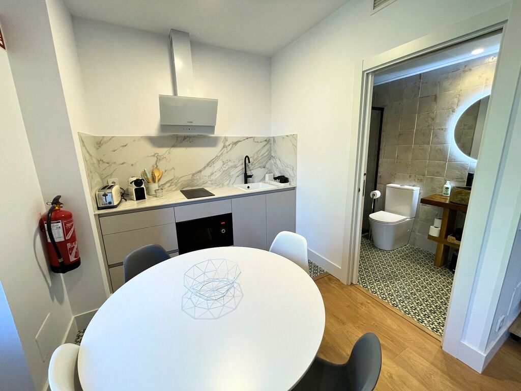 Een compacte keuken met een marmeren achterwand, een ingebouwde oven en een gootsteen. Er staat een eettafel met stoelen, een brandblusser, en er is een kleine badkamer zichtbaar door een geopende deur.