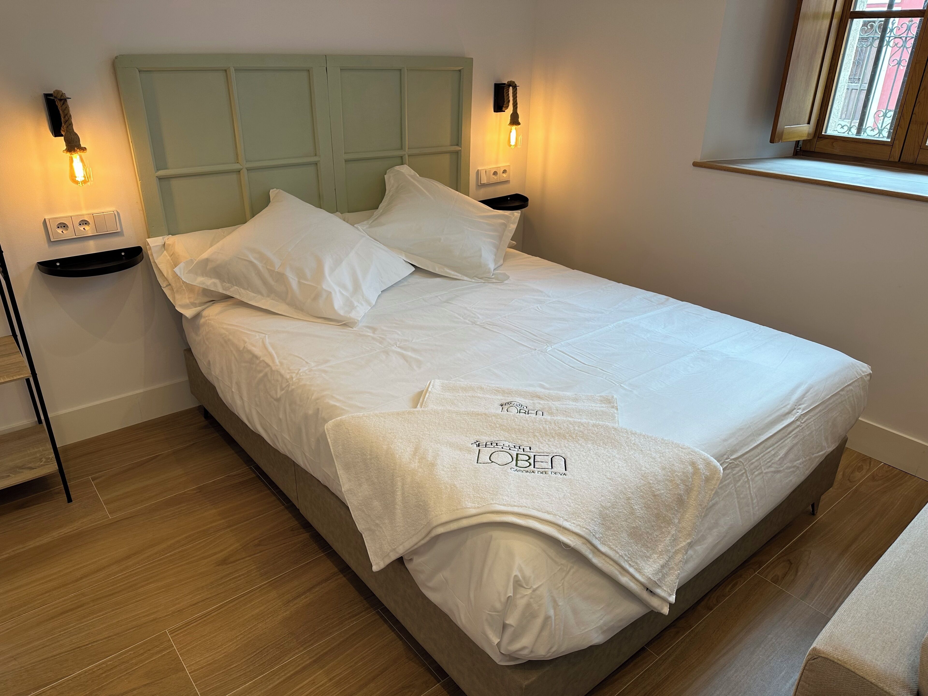 A neatly made bed with white linens and two towels with text, a light green headboard, bedside lamps, and a window with a view.