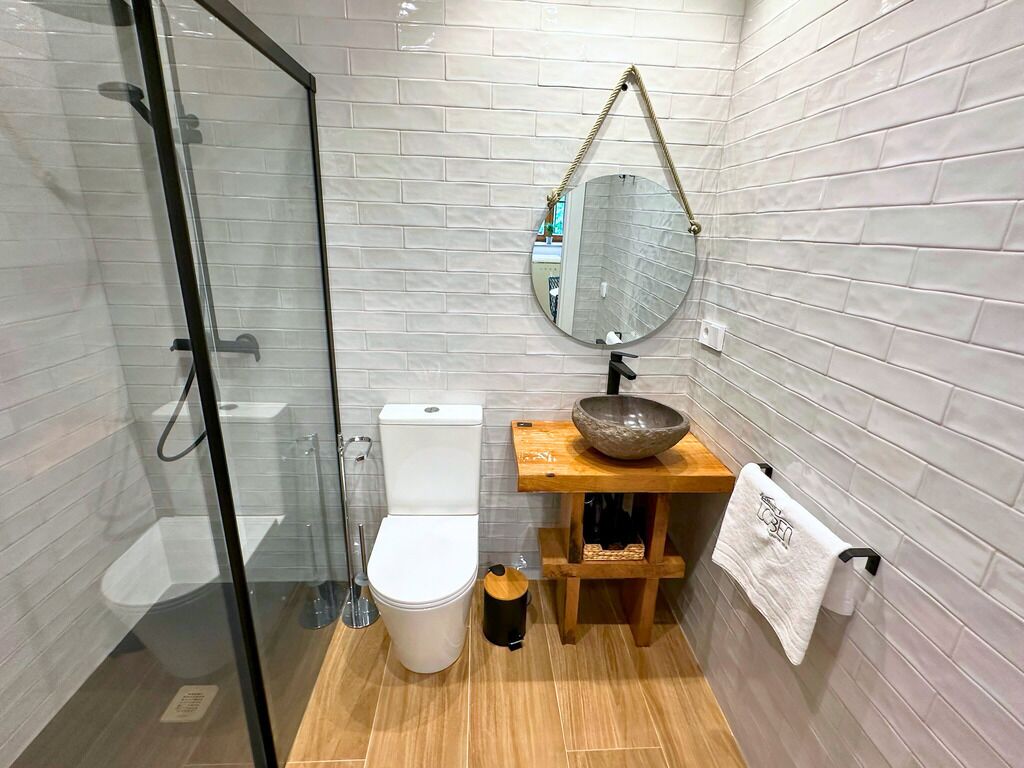 Une salle de bain moderne dotée d’une douche à parois de verre, de toilettes blanches, d’un meuble-lavabo en bois avec un lavabo en forme de coupe et d’un miroir avec une poignée en corde.