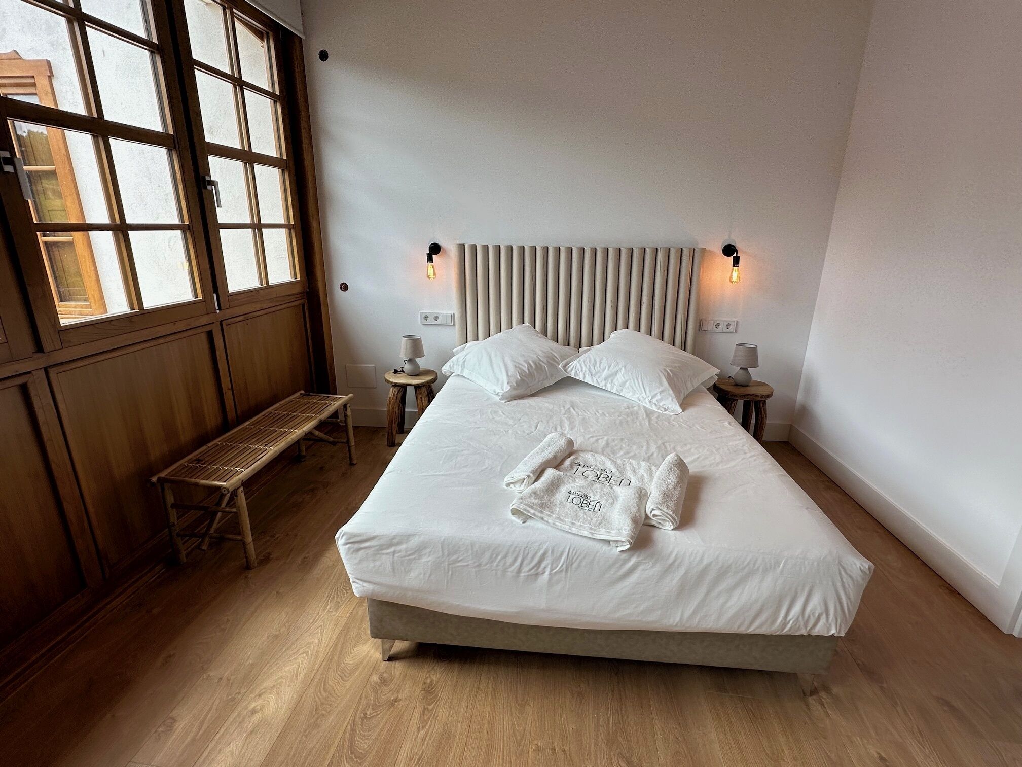 A bedroom with a large bed, two bedside tables, a bench, and a window with blinds.