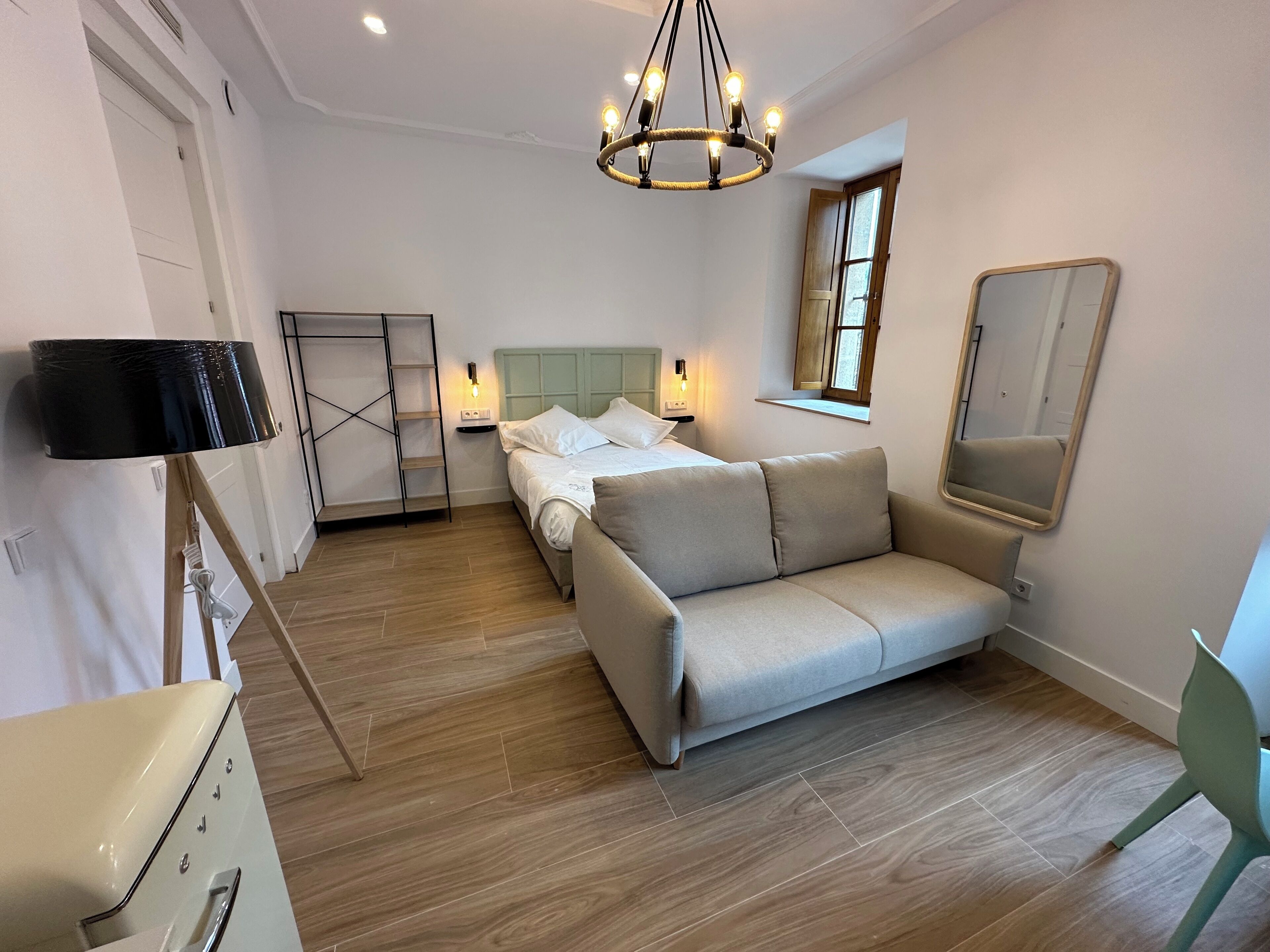 A bedroom with a bed, a sofa, a desk, a mirror, and a chandelier.