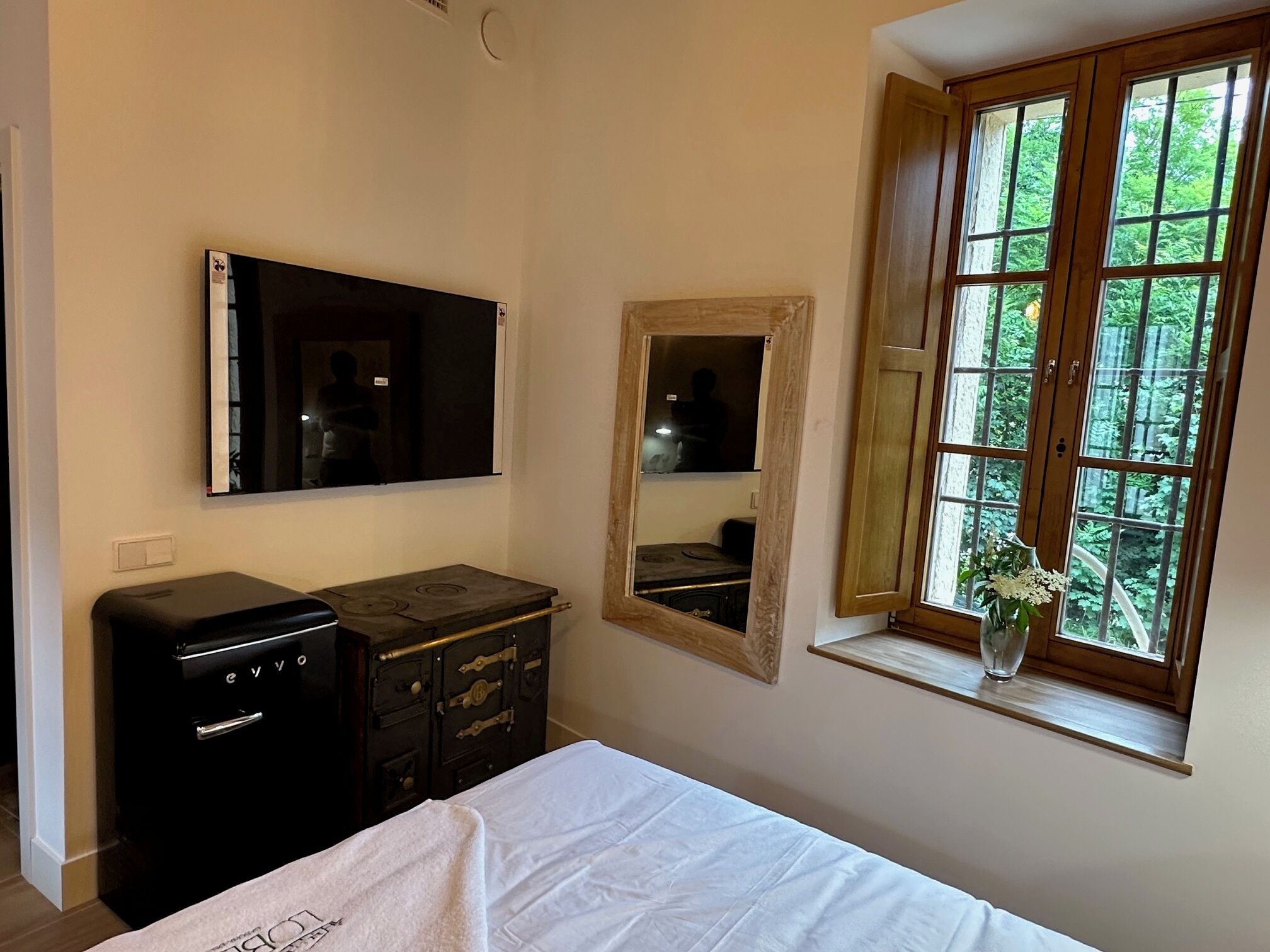 Een slaapkamer met een bed, een dressoir, een televisie, een spiegel en een raam met uitzicht op groen.
