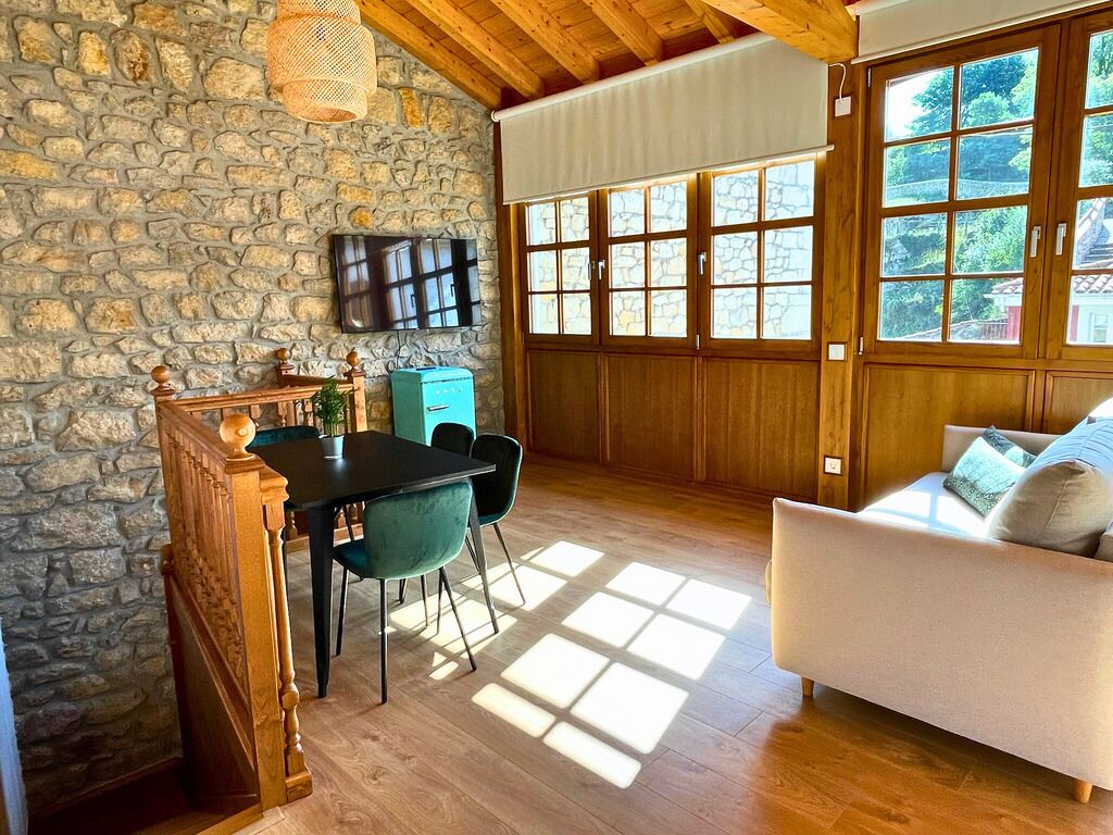 A cozy room with a stone wall, wooden ceiling, a dining table with chairs, a sofa, and large windows with a view of greenery.