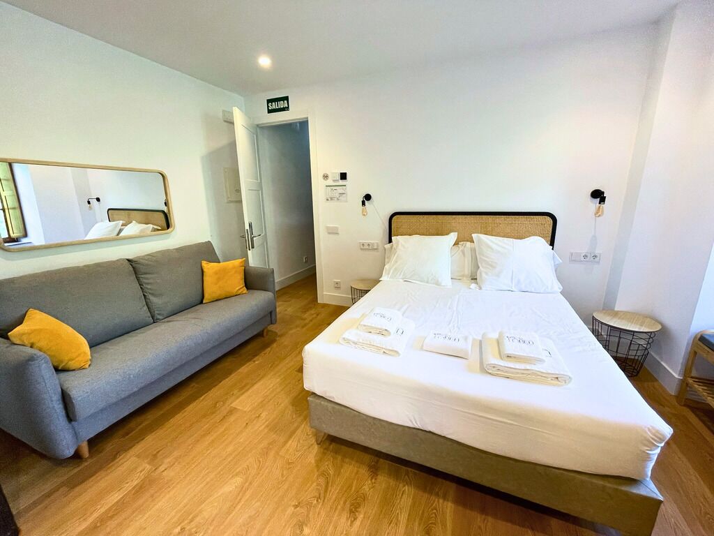 Une chambre d’hôtel avec un lit, un canapé gris agrémenté de coussins jaunes, un miroir et une porte portant l’inscription « SORTIE ».