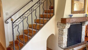 Una escalera con barandillas metálicas decoradas y peldaños de madera, que conduce a una chimenea de piedra con repisa y protector.