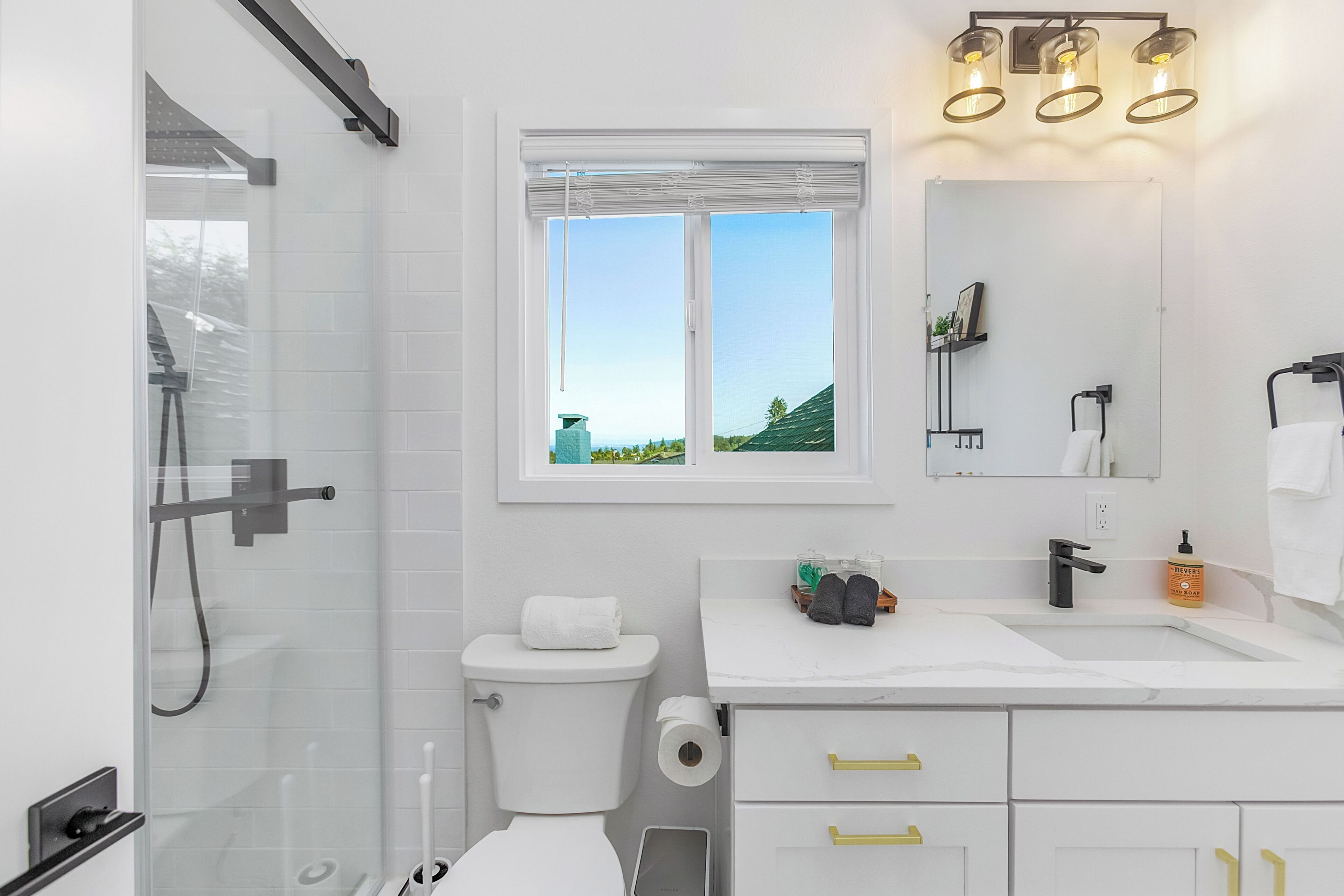 A modern bathroom with a glass shower enclosure, a toilet, a sink with a mirror, and a window offering a view of greenery.