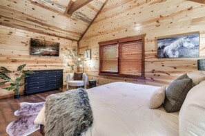 A log cabin interior with a bed, a dresser, a chair, and a window with blinds.