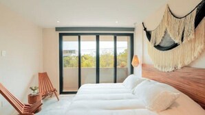 Un dormitorio con una cama grande, dos sillas de madera, un colgador de macramé y vistas a través de un ventanal corredero.