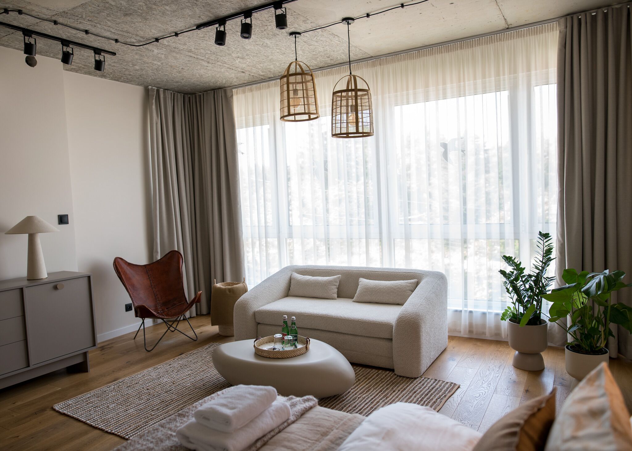 A modern living room with a sofa, a chair, a coffee table, and a lamp. There are curtains, a plant, and a window with natural light.