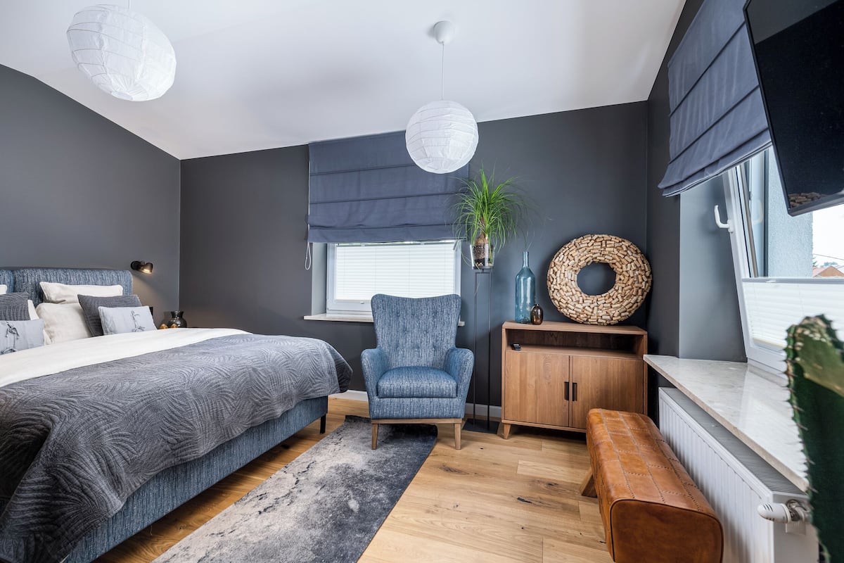 A modern bedroom with a large bed, a blue armchair, a wooden bench, and a window with a view.