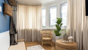 Una habitación con una silla de mimbre, un otoman redondo, un tapete tejido y una ventana con cortinas.