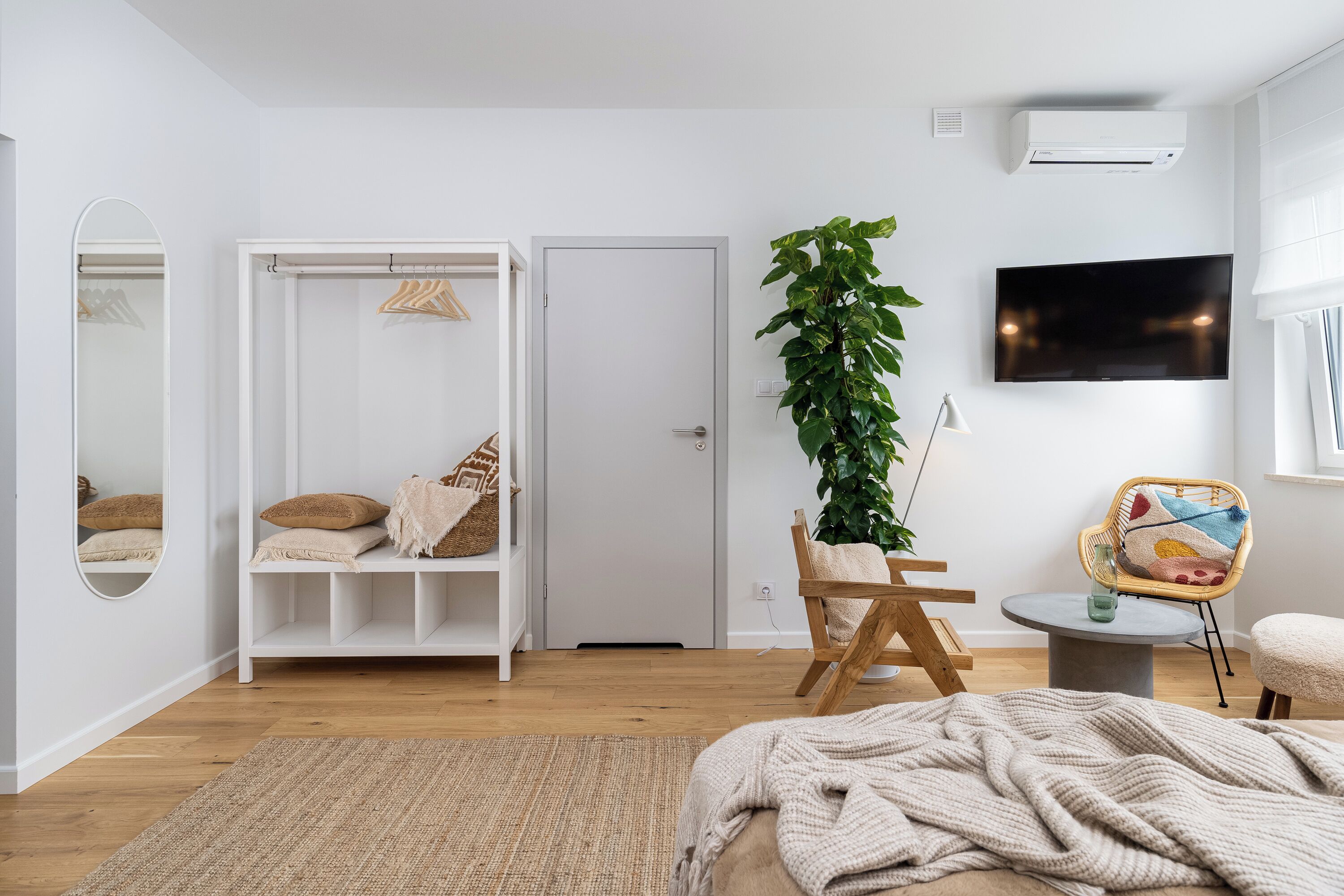 Een moderne slaapkamer met een bed, een kledingkast, een spiegel, een plant, een televisie en een klein rond tafeltje.