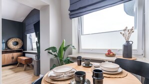 Un comedor moderno con una mesa redonda dispuesta para dos personas, con platos, tazas y un jarrón de flores secas.