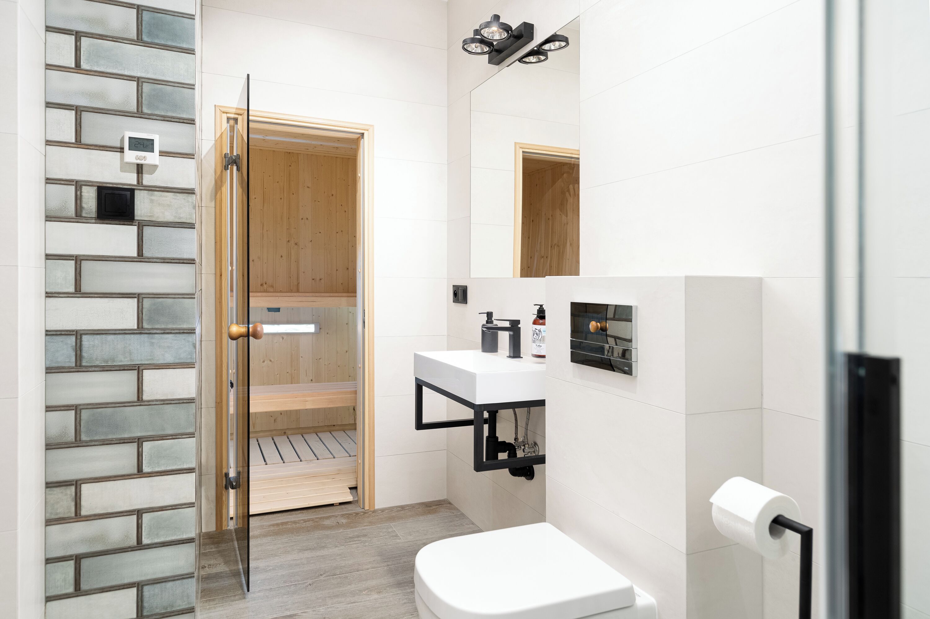 Un bagno moderno con box doccia in vetro, un water bianco, un lavandino con rubinetto nero e una porta in legno che dà accesso alla zona doccia.