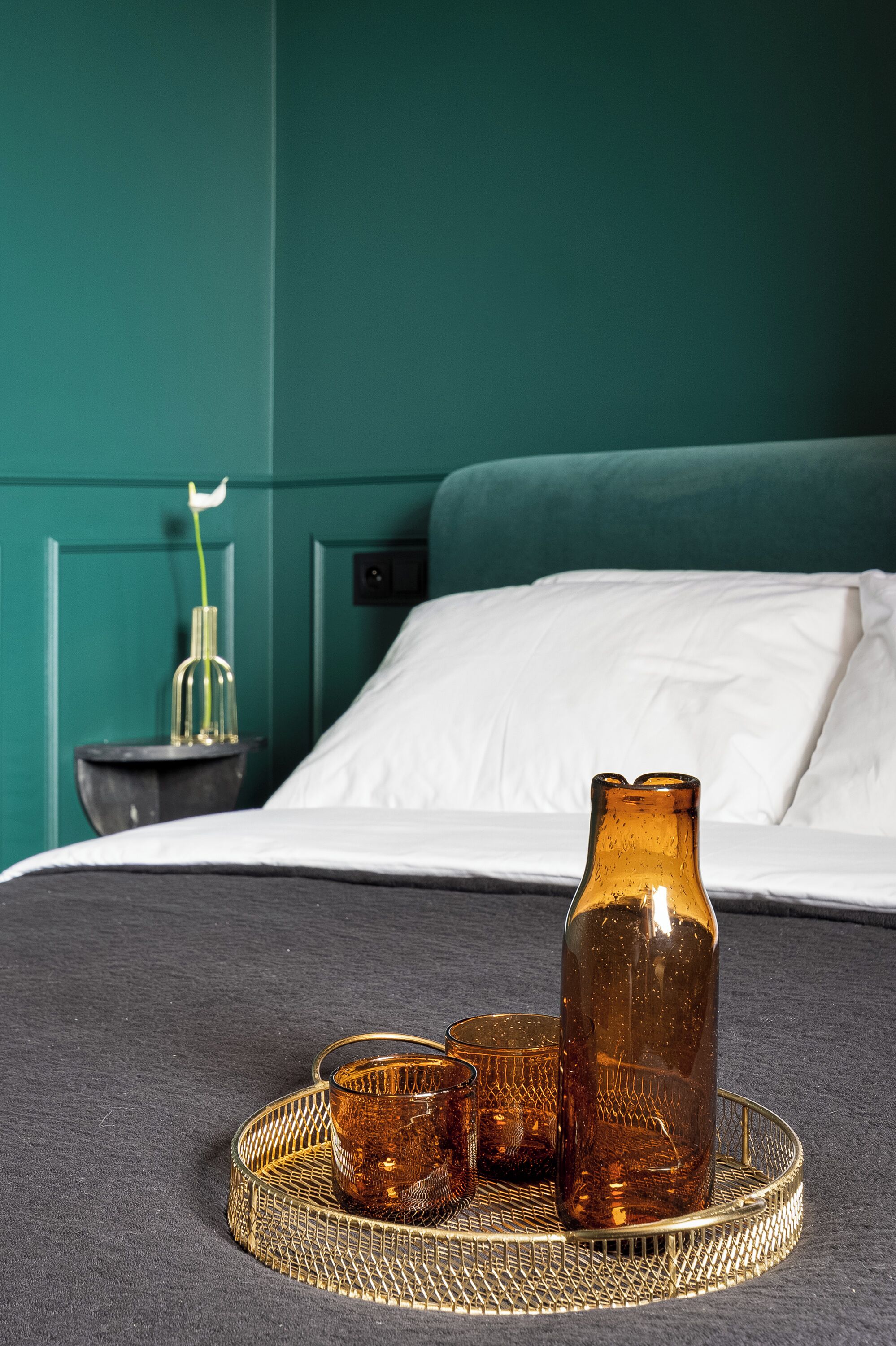 A bed with a white pillow and a teal headboard, a bedside table with a vase and a small plant, and a tray with a glass and a bottle.