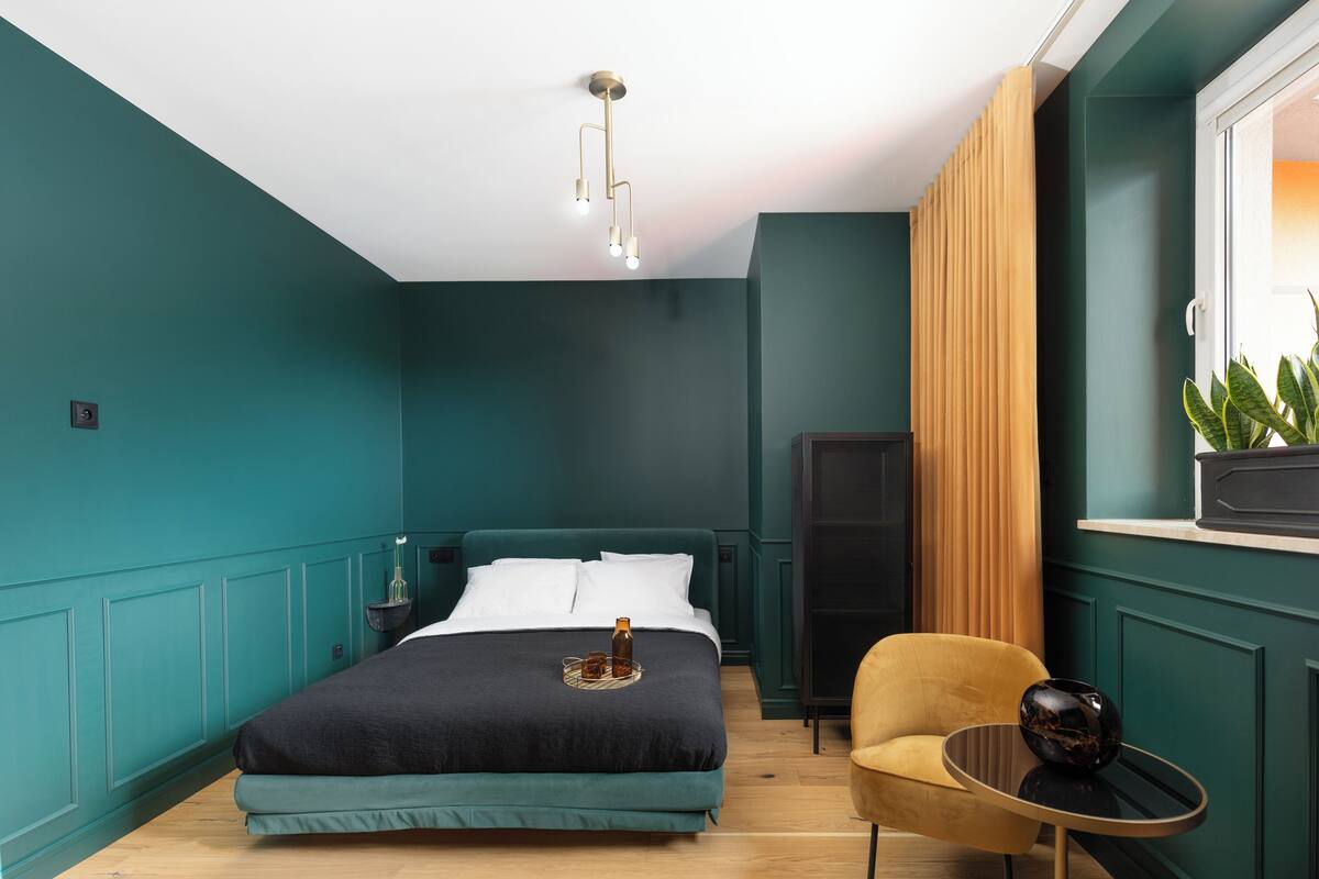 A modern bedroom with teal walls, a bed with a dark cover, a bedside table with a bottle and glasses, a yellow armchair, and a window with a plant.