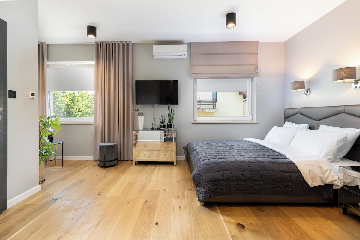 A modern bedroom with a large bed, a flat-screen TV, a bedside table, a dresser, and a window with curtains.